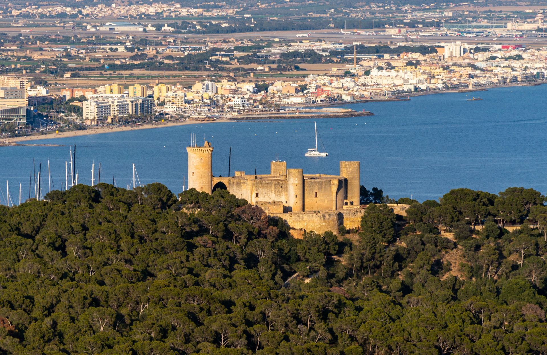 Stone castle on a wooded hill overlooking a blue bay and distant city skyline.