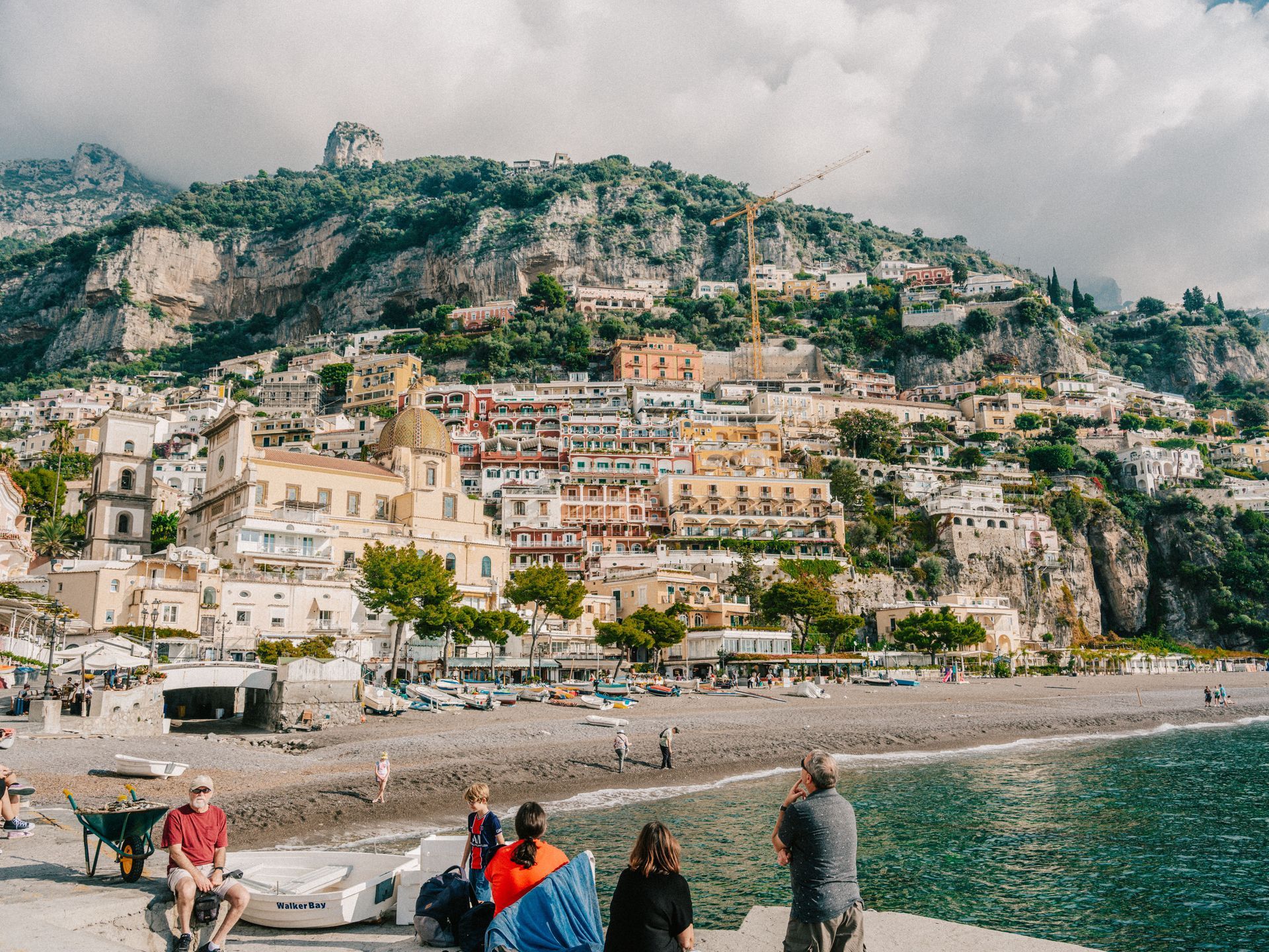 Crowded seaside beach beneath a cliffside town with pastel buildings and mountains in the background