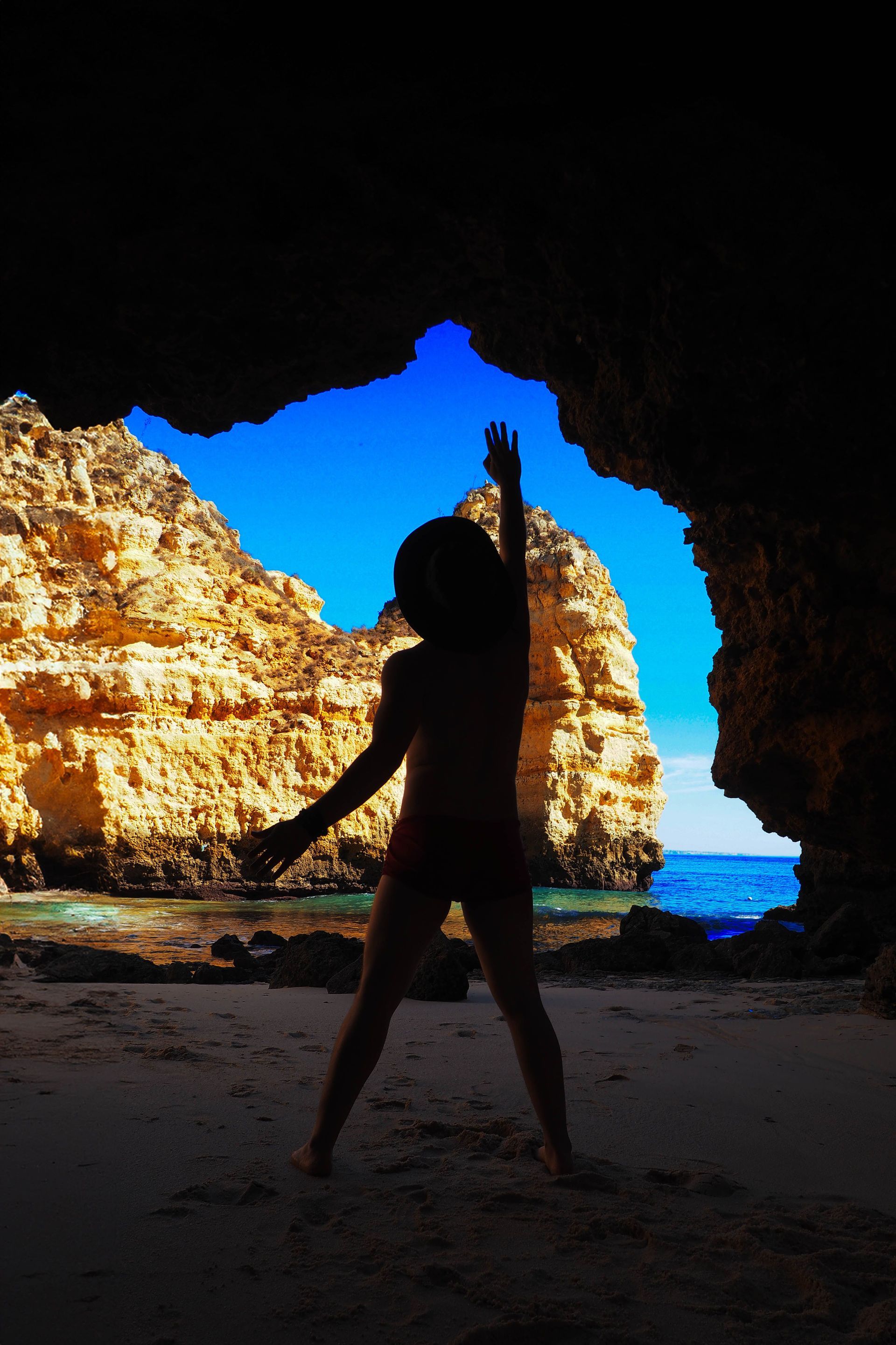 Silhouetted person standing in a cave opening, with bright blue sky and ocean beyond.