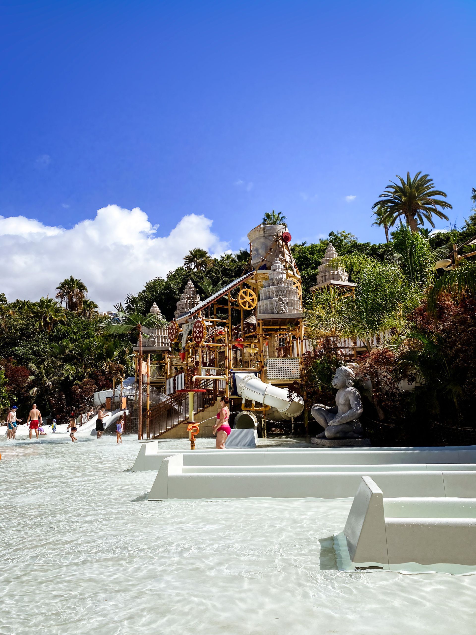 Tropical resort pool with people, white steps, and a colorful slide under a blue sky