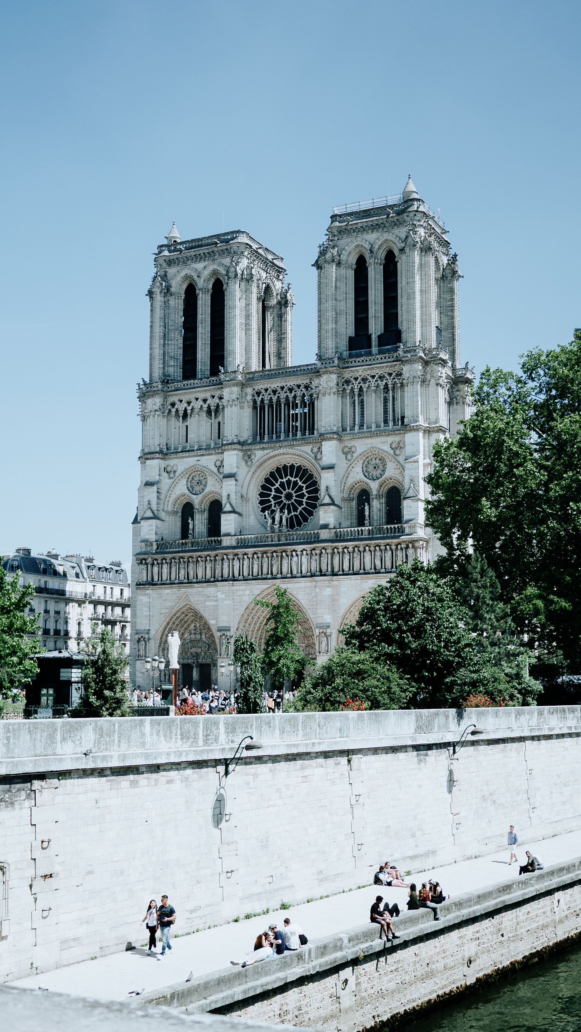 Notre-Dame Cathedral behind a riverside wall under a clear blue sky, with a few people below