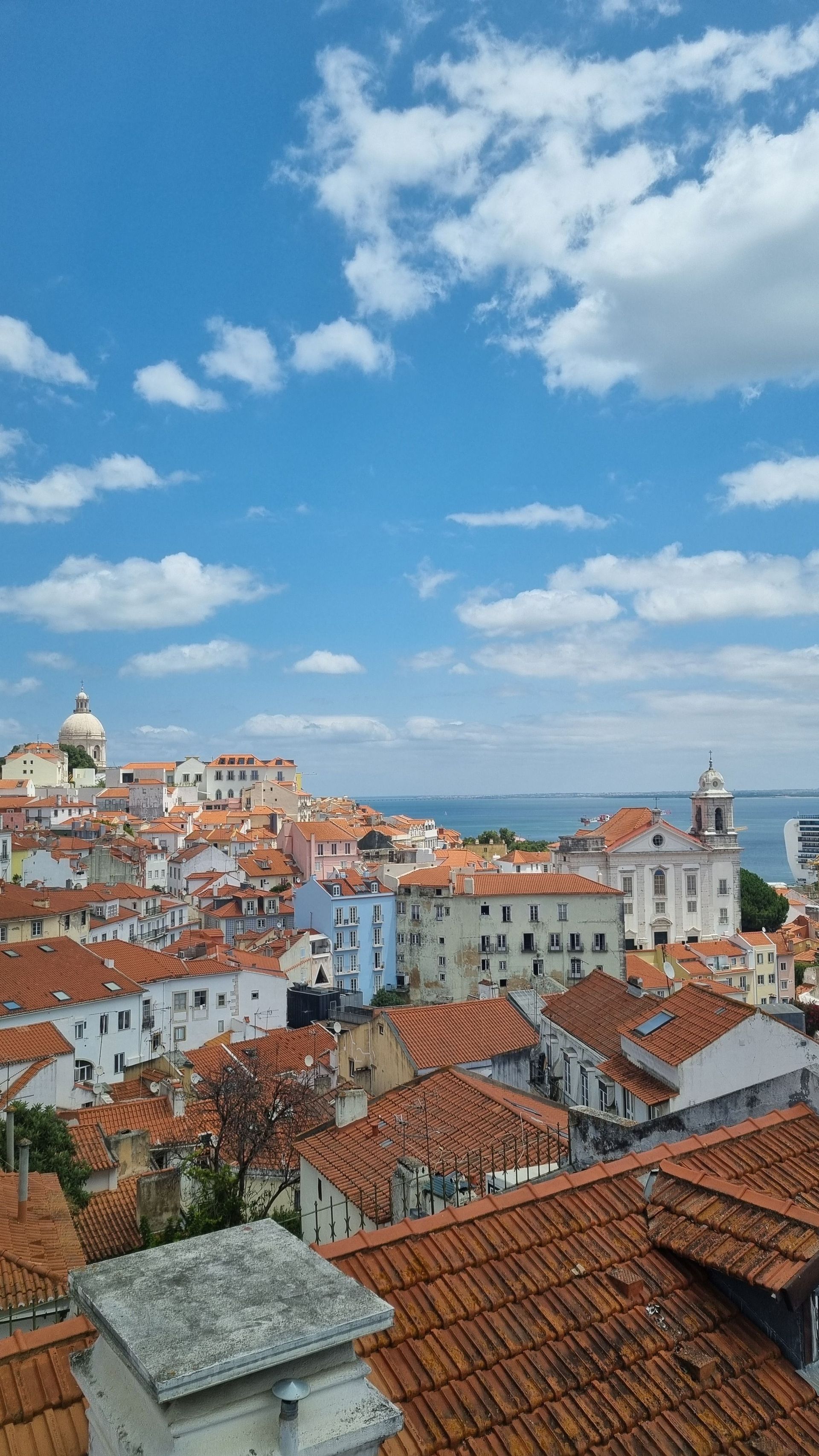 Red-roofed coastal city skyline under a bright blue sky with scattered clouds