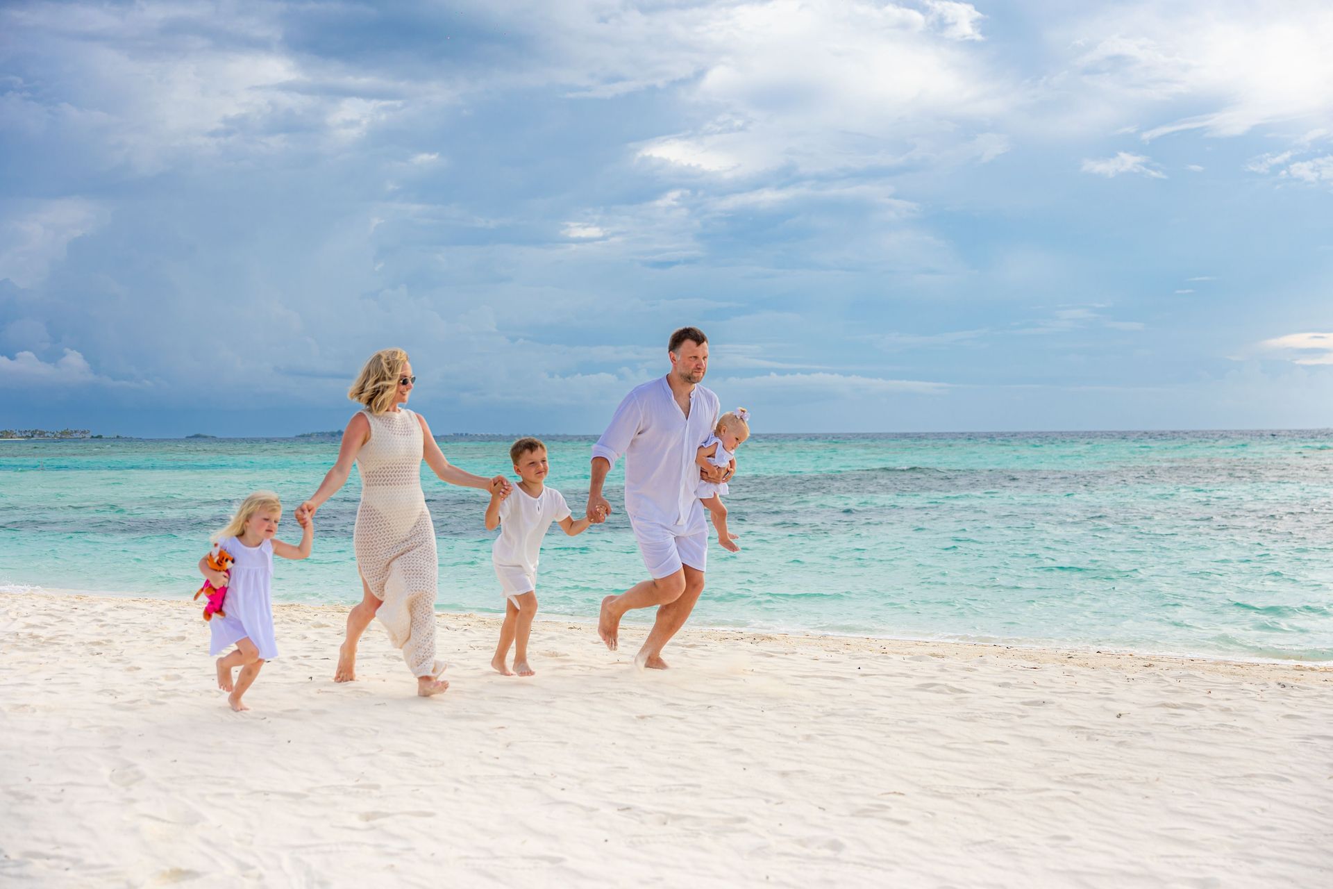 Family running hand-in-hand along a bright white beach beside turquoise water under a cloudy sky