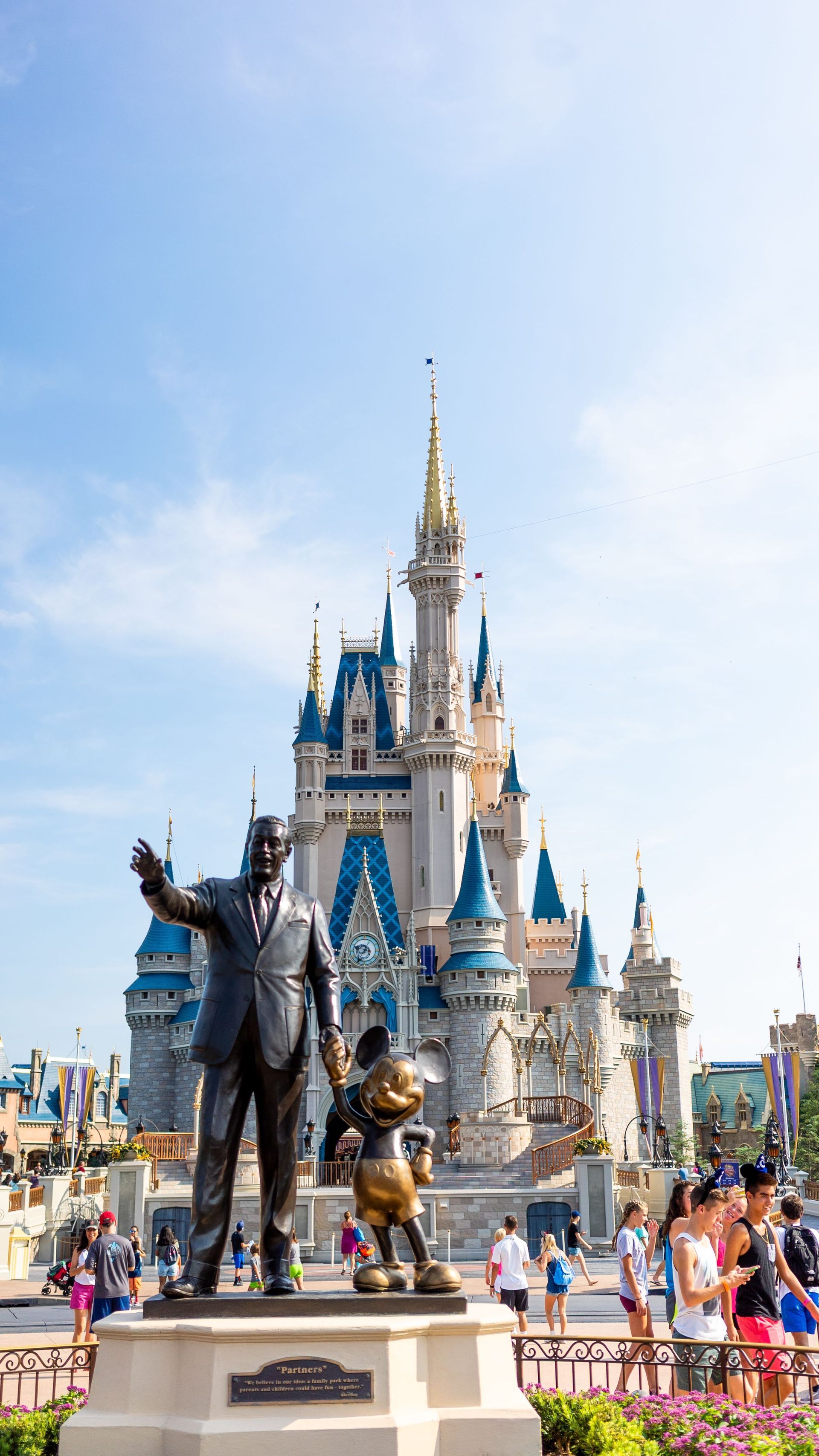 Cinderella Castle at Disney, with Mickey and Minnie statues in front and visitors gathered nearby under a blue sky
