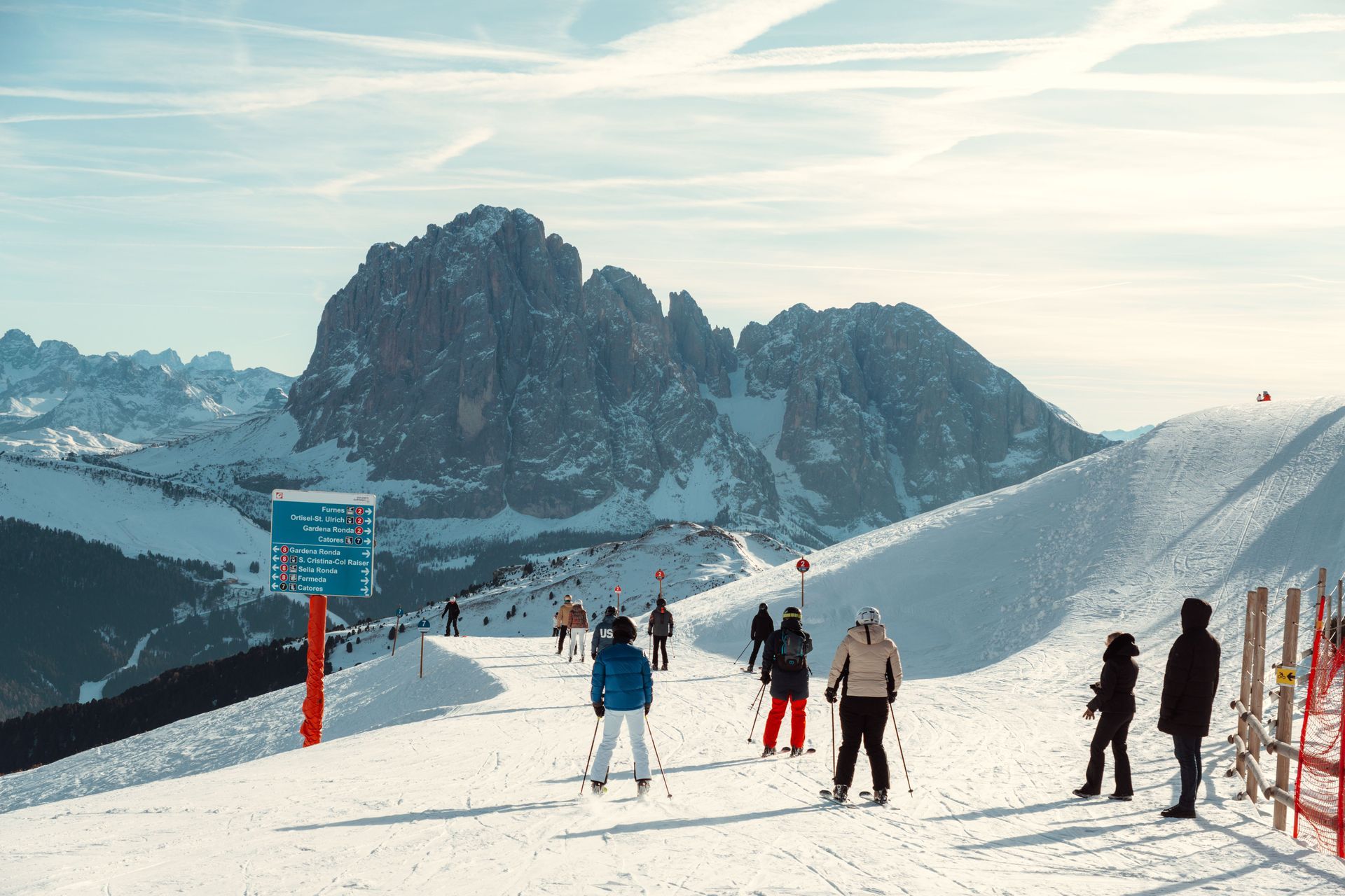 Skiers standing on a snowy mountain slope with rugged peaks and a blue trail sign under a clear sky