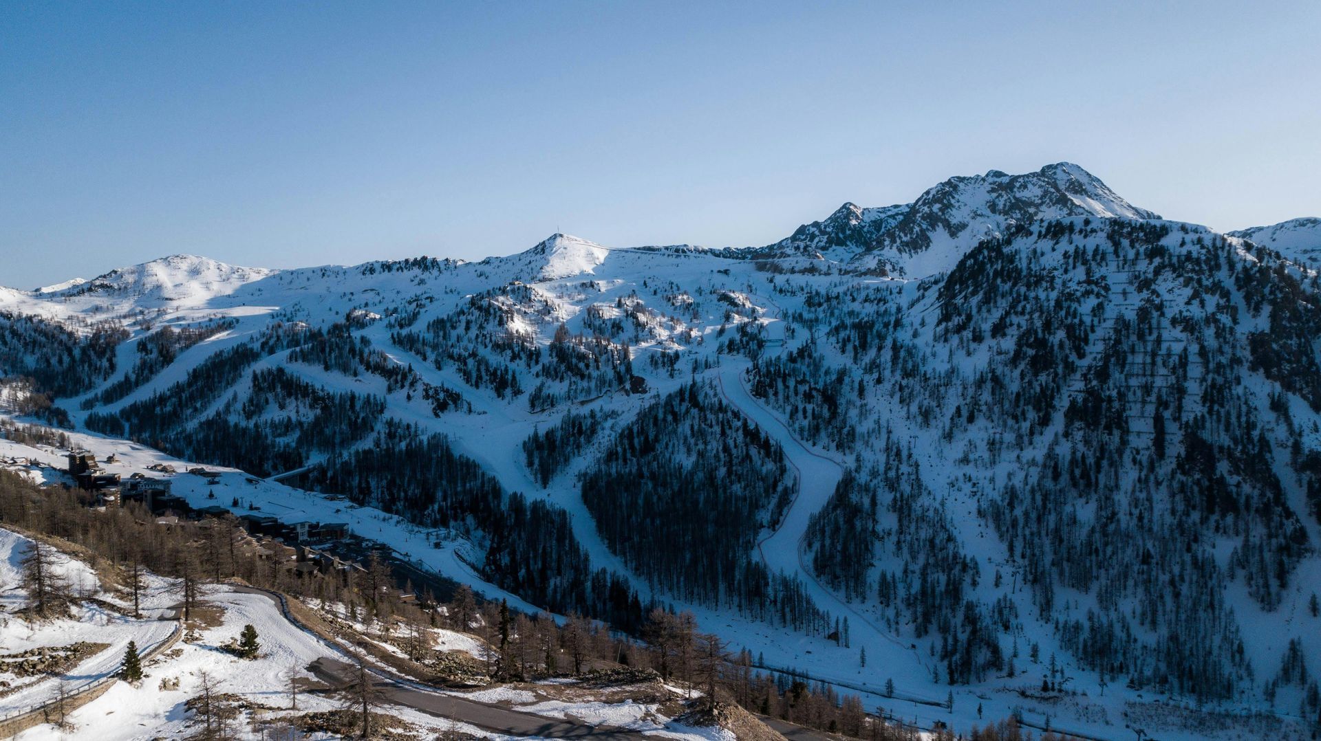 Snow-covered mountain range with steep, forested slopes under a clear blue sky