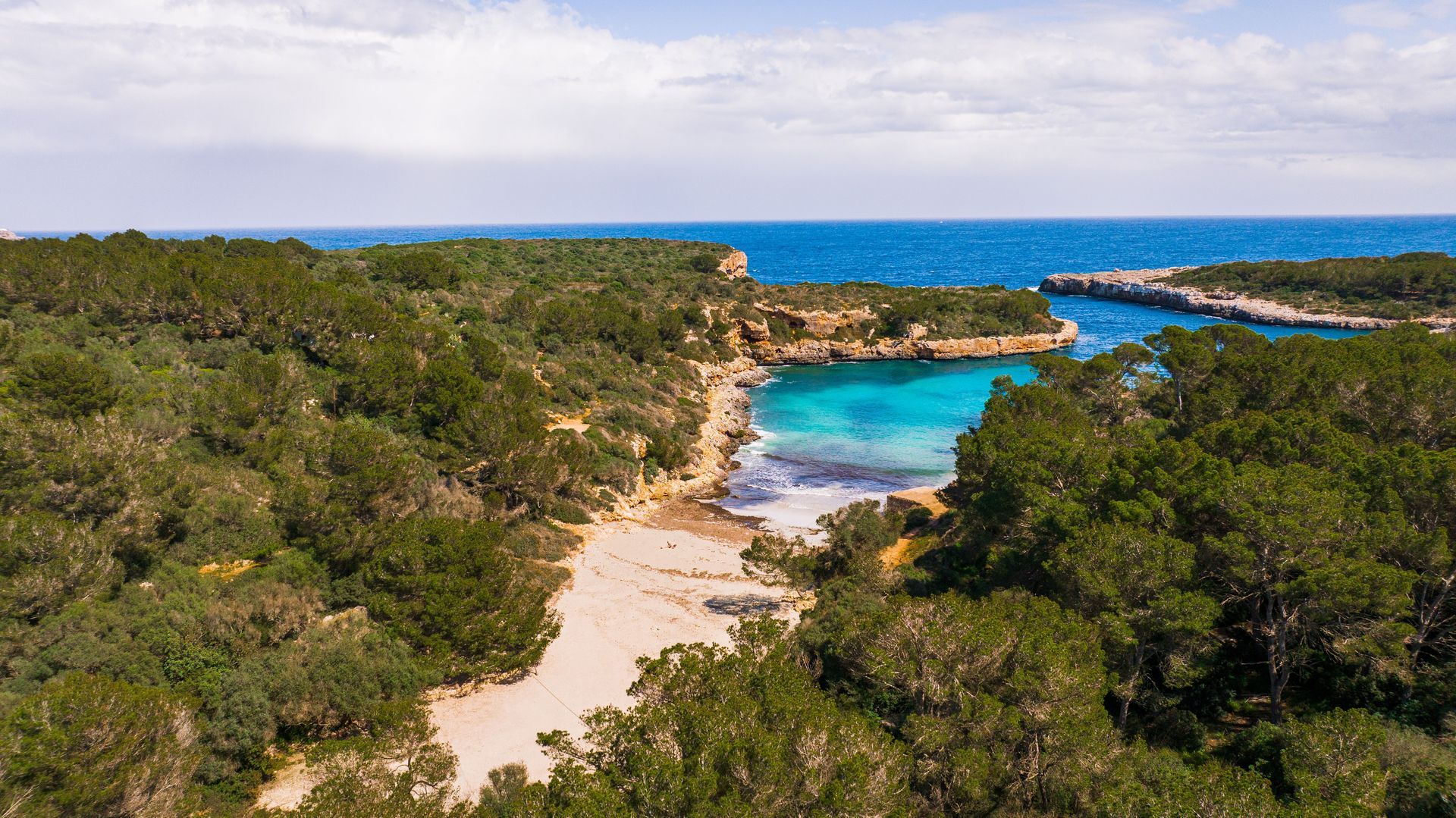 Tropical shoreline with turquoise water, rocky coast, sandy beach, and dense green vegetation under a cloudy sky