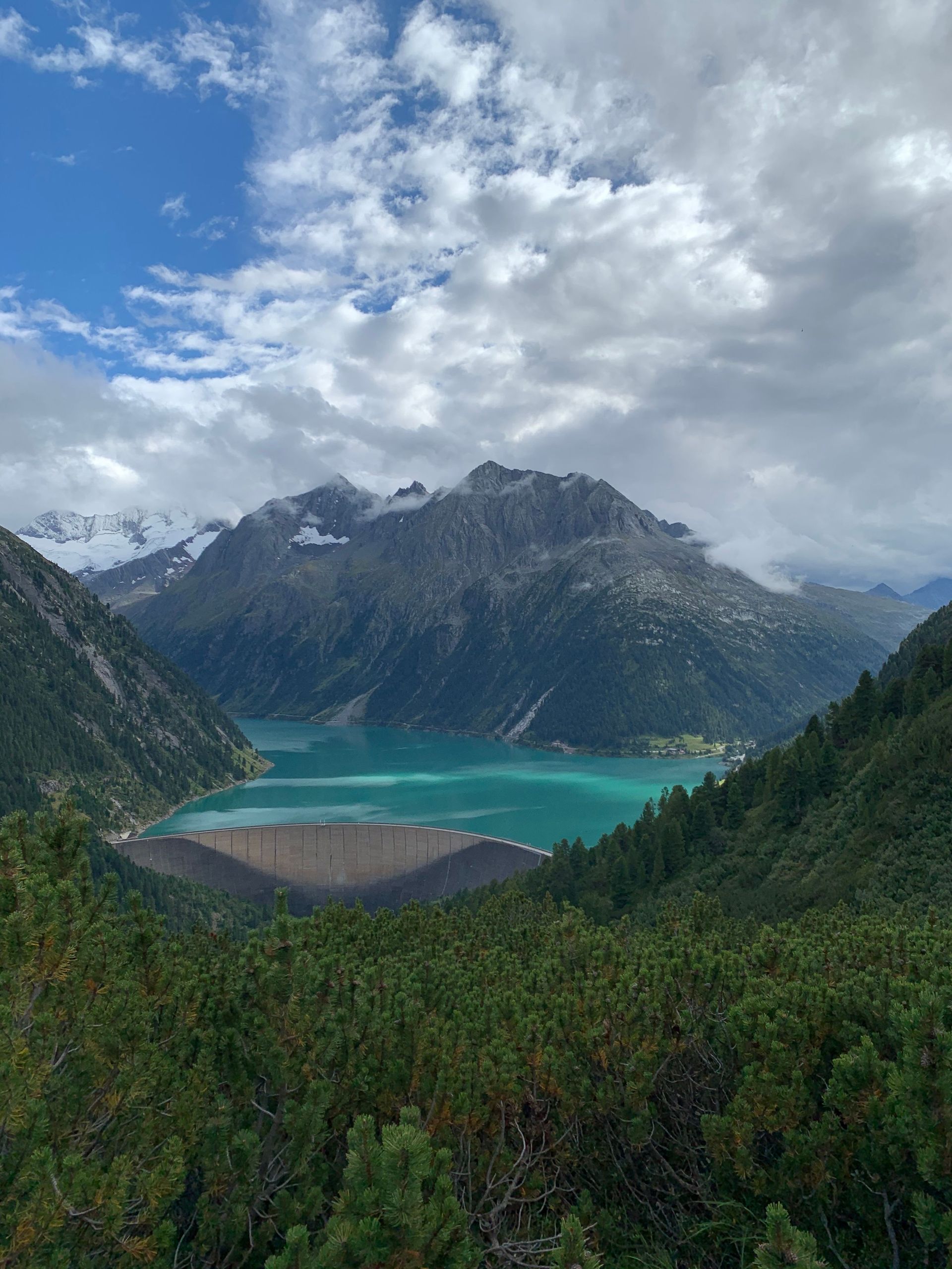 Turquoise mountain lake beneath snowy peaks and cloudy sky, with forested foreground