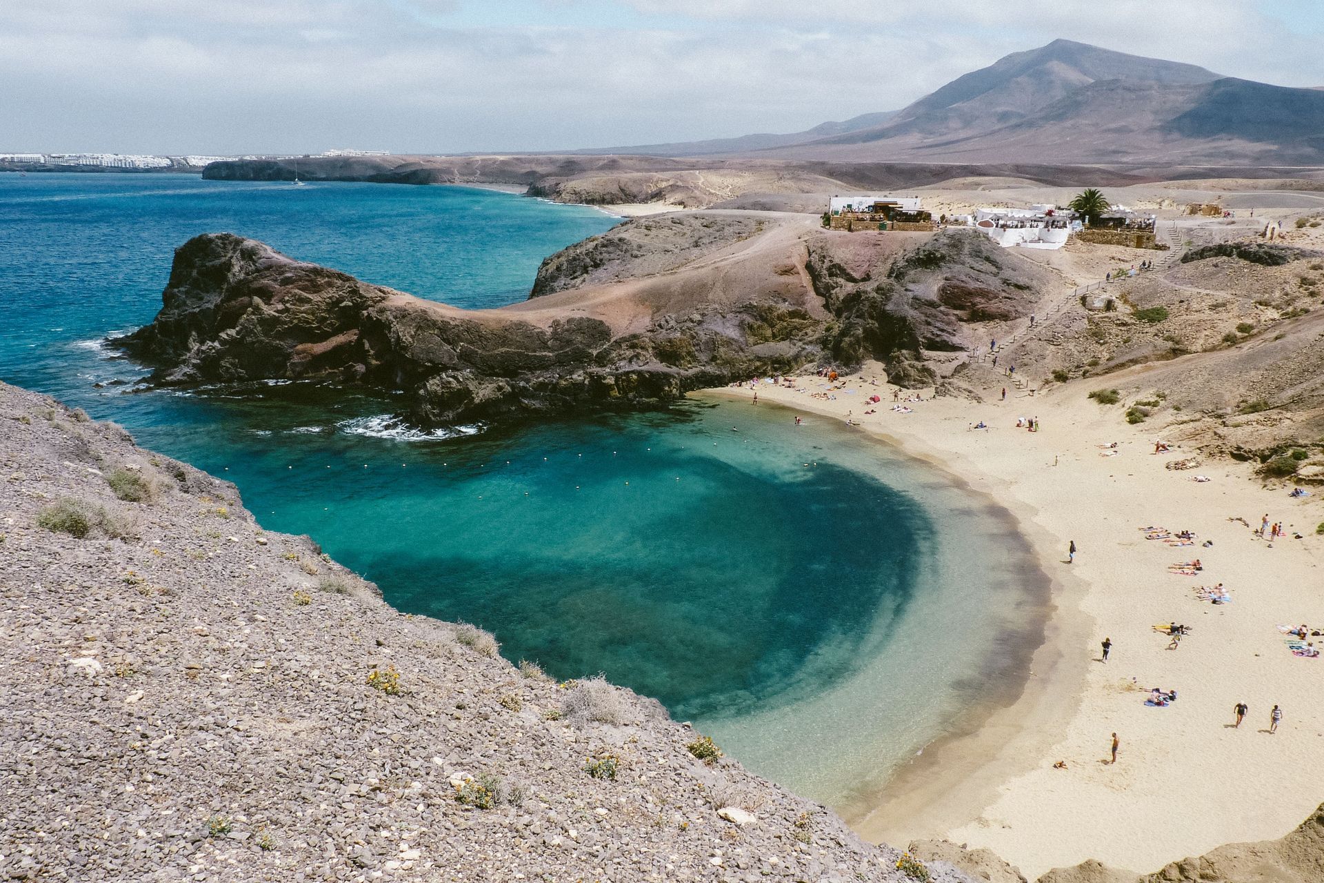 Turquoise bay with sandy beach, rocky shore, and distant mountains under a clear sky