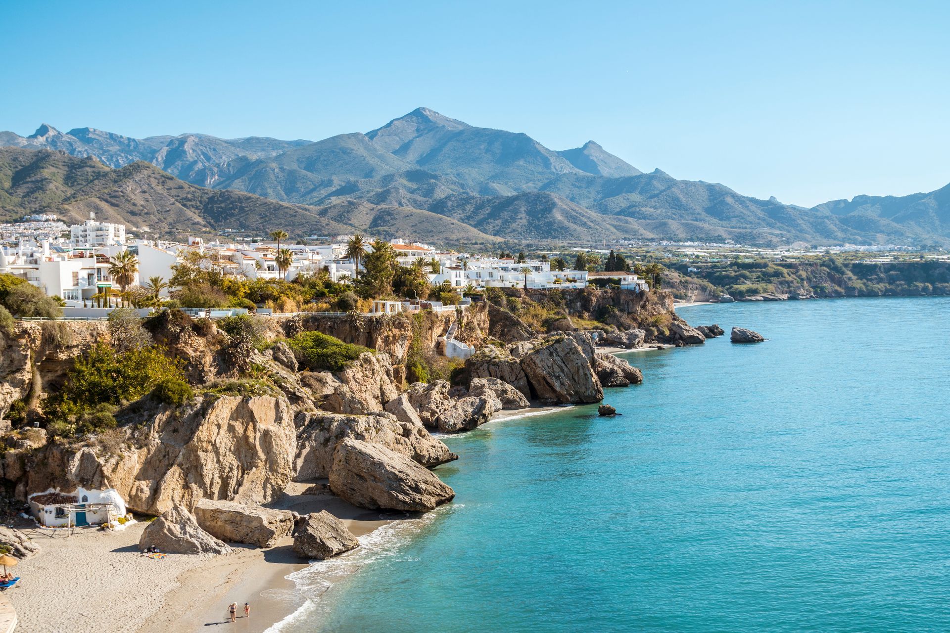 Cliffside seaside town with white buildings, beach, and turquoise water beneath mountains
