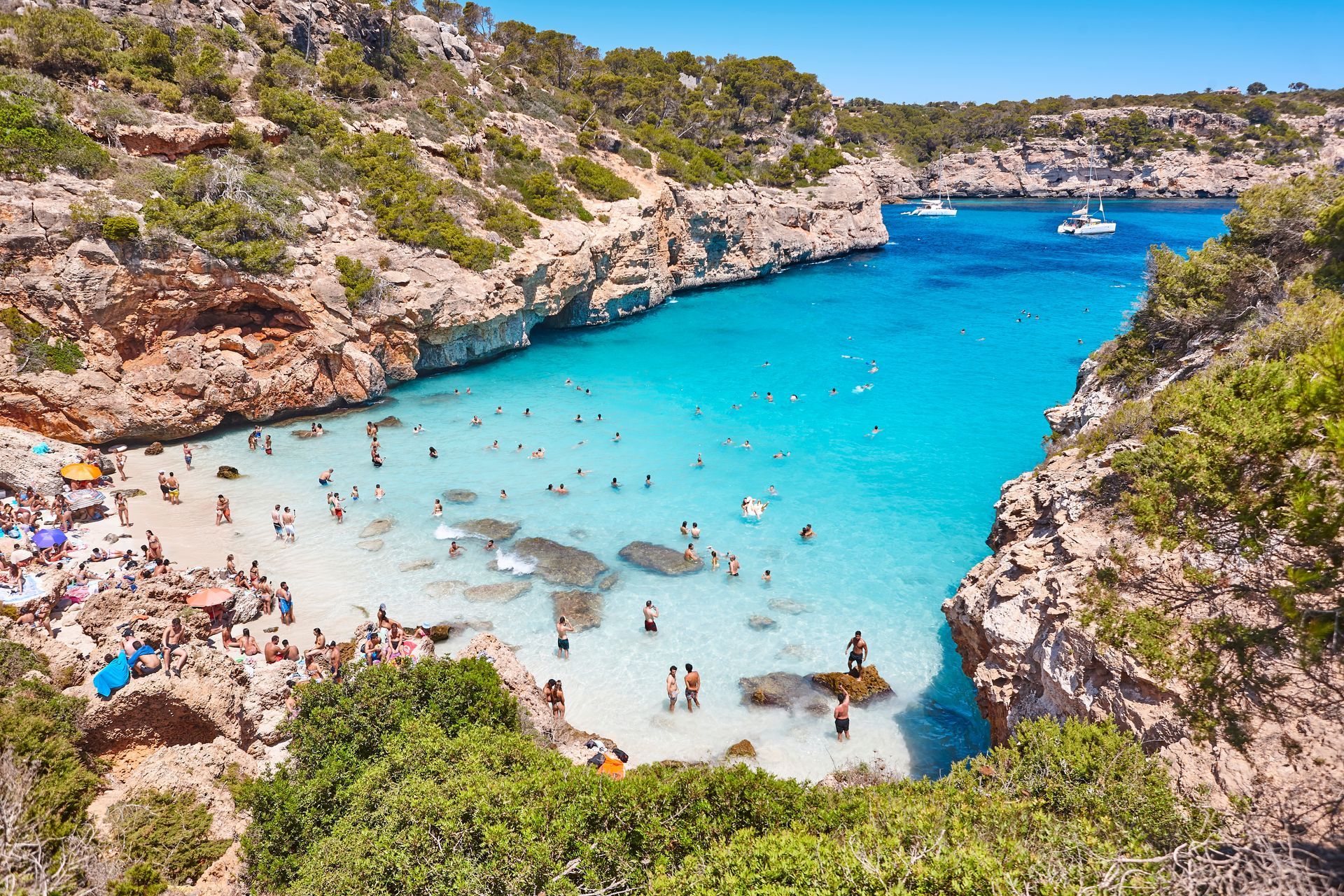 Crowded turquoise cove beach with swimmers, rocky cliffs, and small boats under a sunny sky
