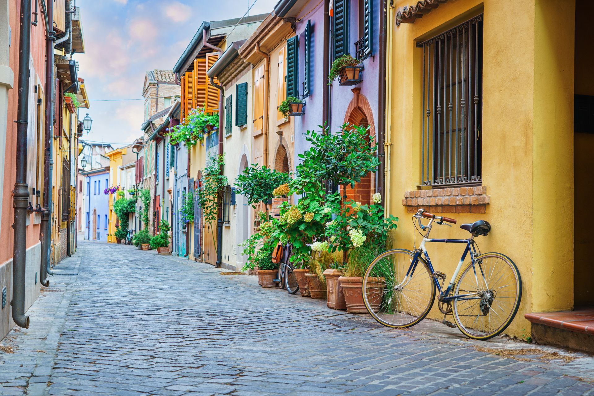 Colorful cobblestone street with pastel houses, potted plants, and a bicycle leaning against a yellow wall.