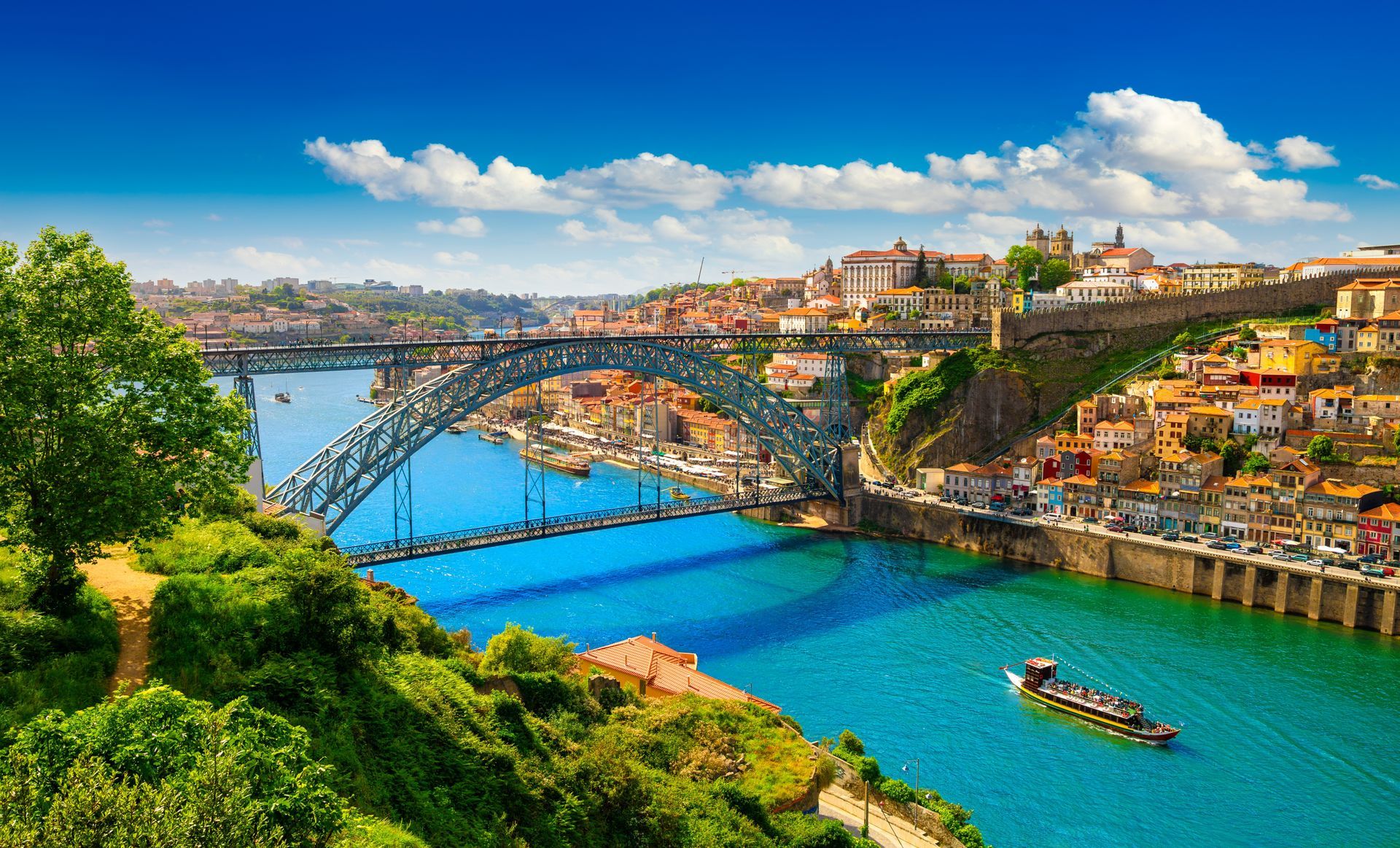 Panoramic view of a river city with a stone bridge, colorful hillside buildings, and boats on blue water