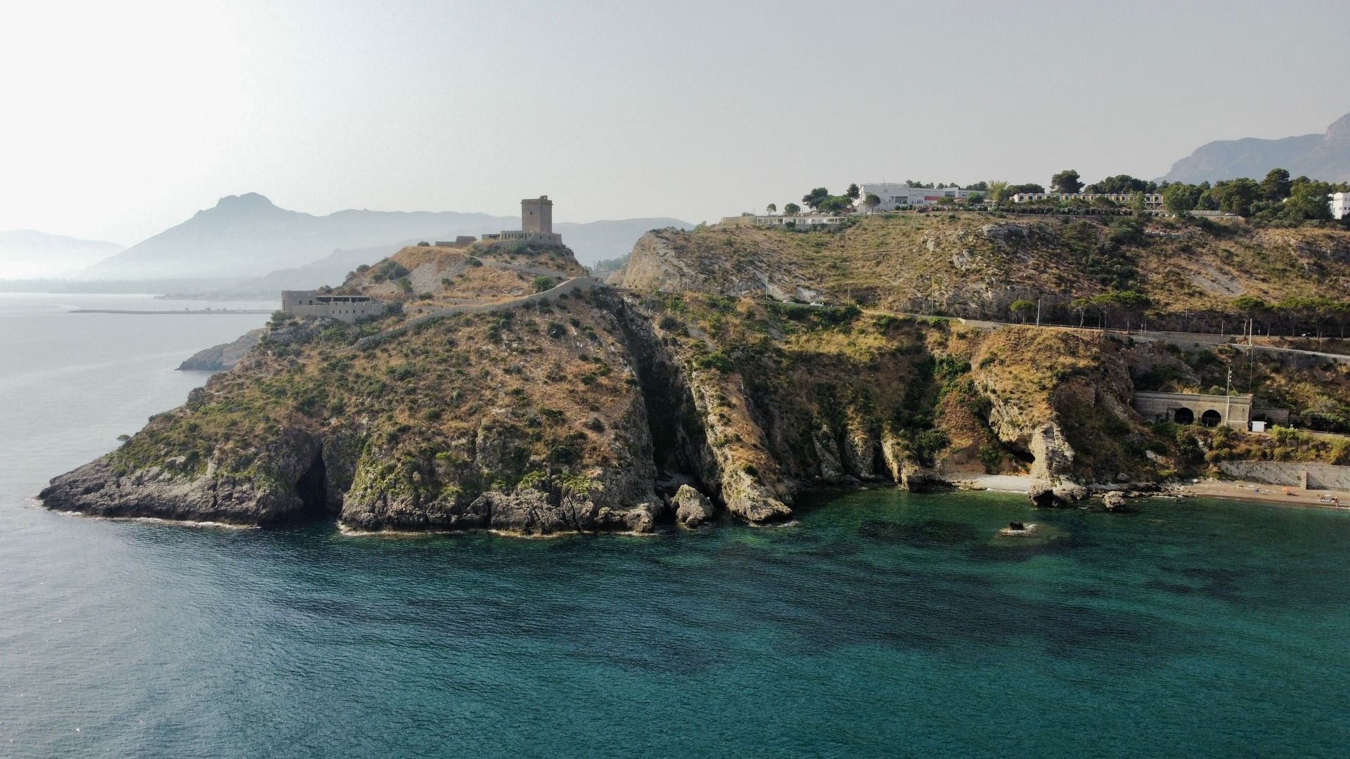 Rocky coastal cliff with a small fort overlooking calm blue water and distant misty mountains