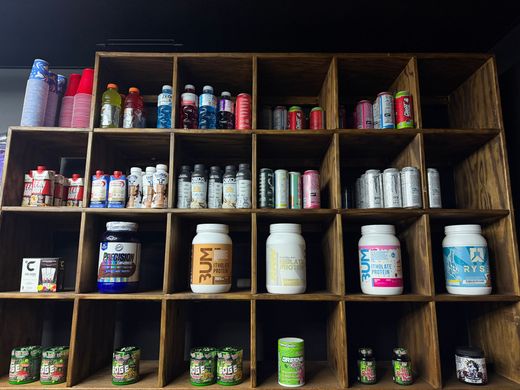 Wooden shelf stocked with assorted protein and supplement canisters, bottles, and small packaged items.