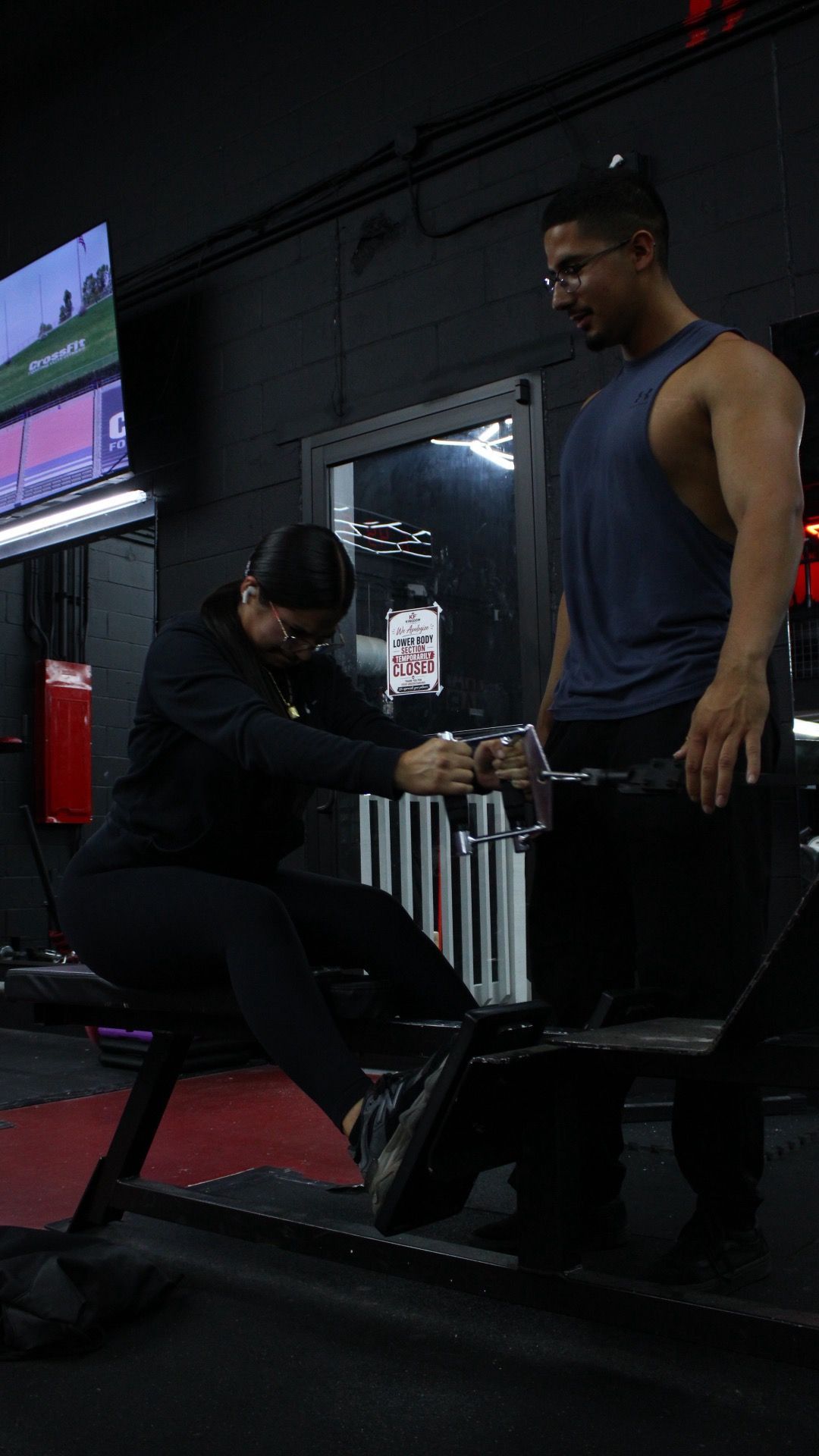 Person doing a cable exercise in a dim gym while a trainer assists nearby