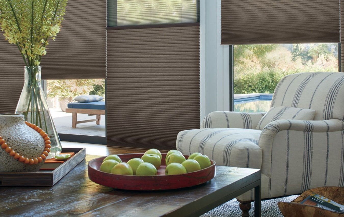 A living room with blinds, a chair and a table with a bowl of apples on it.