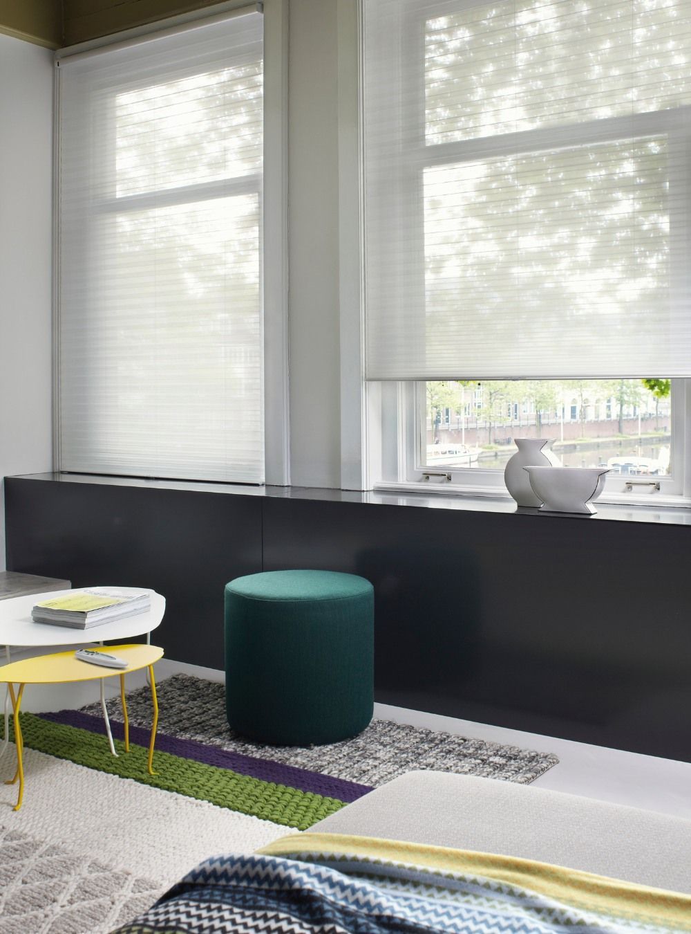 A living room with a table , ottoman , and two windows and blinds