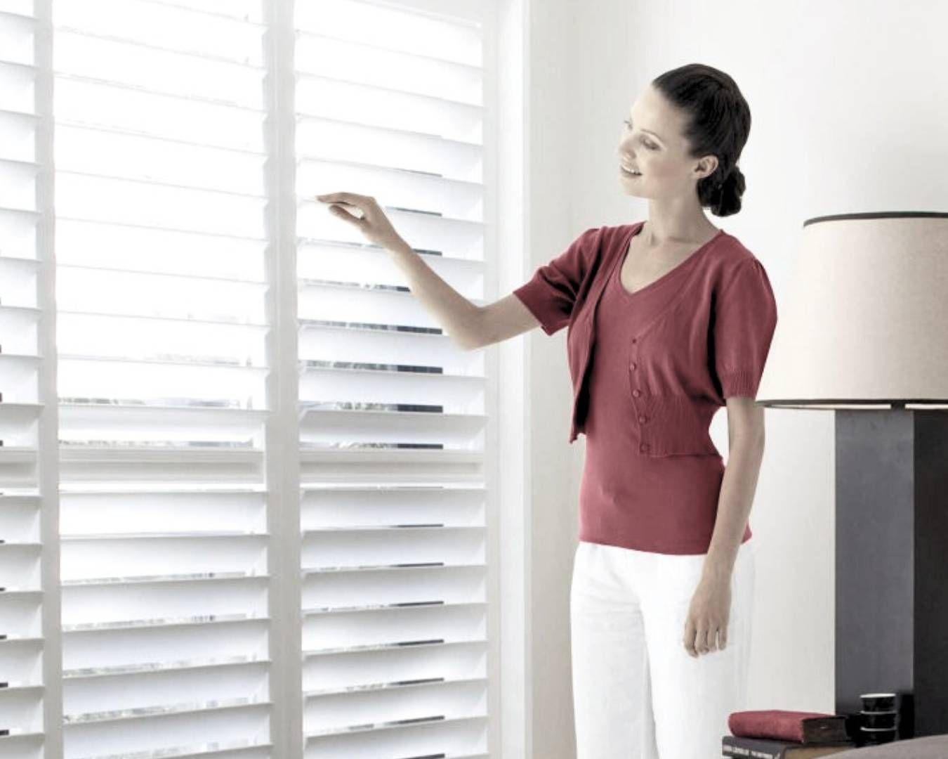 A woman in a red shirt is standing in front of a window with white blinds.