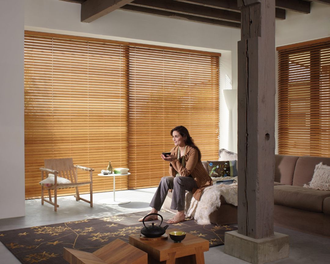 A woman is sitting on a couch in a living room holding a cup of coffee with venetian blinds