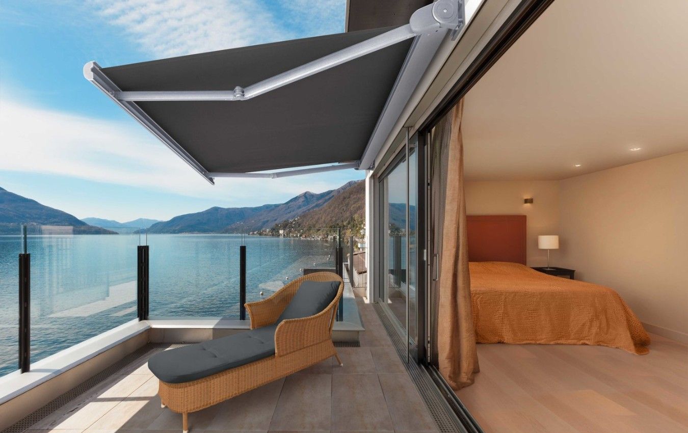 A balcony with a chair, awning and a bed overlooking a lake.