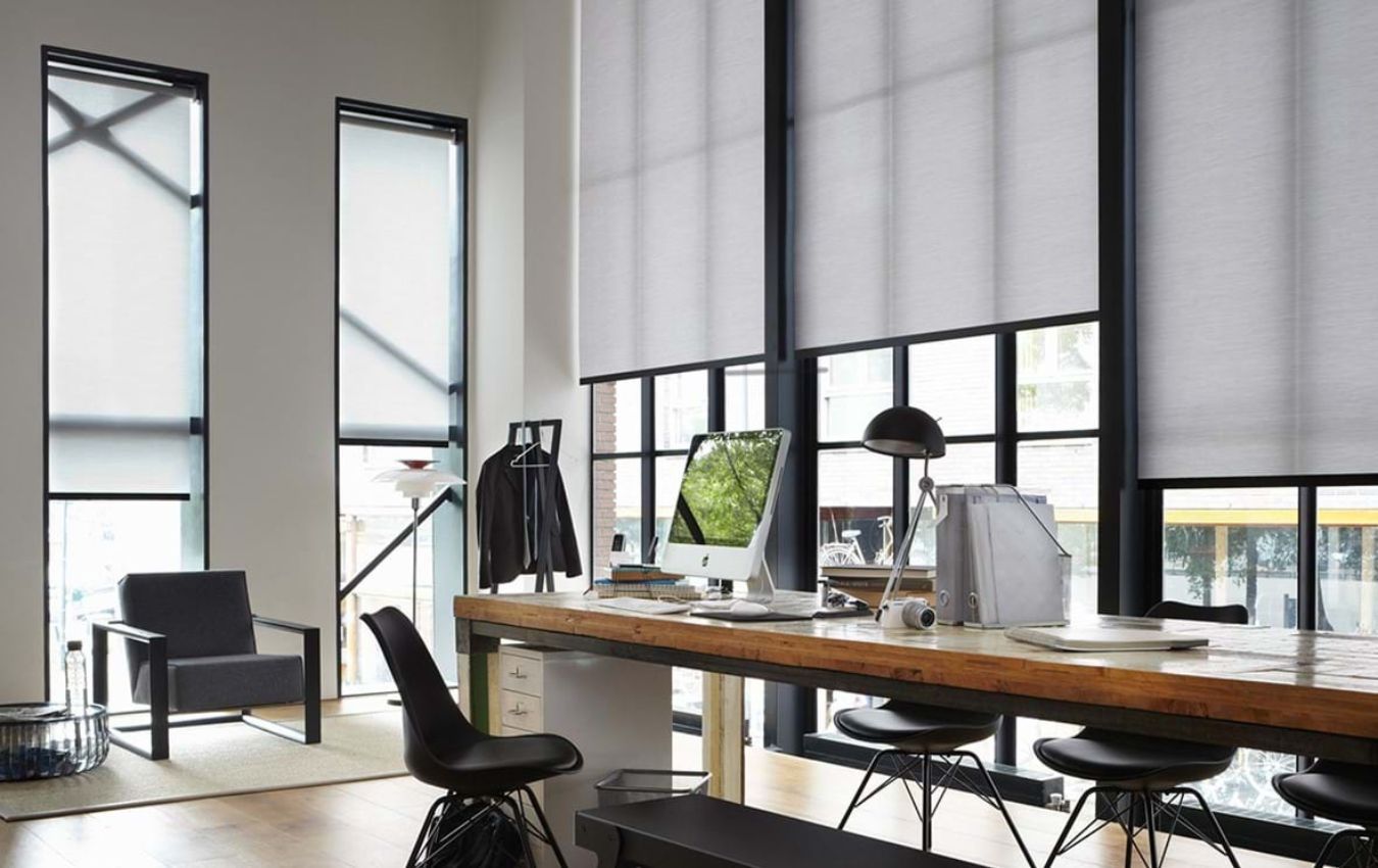 Modern office space with large windows featuring roller blinds, desks, and chairs.