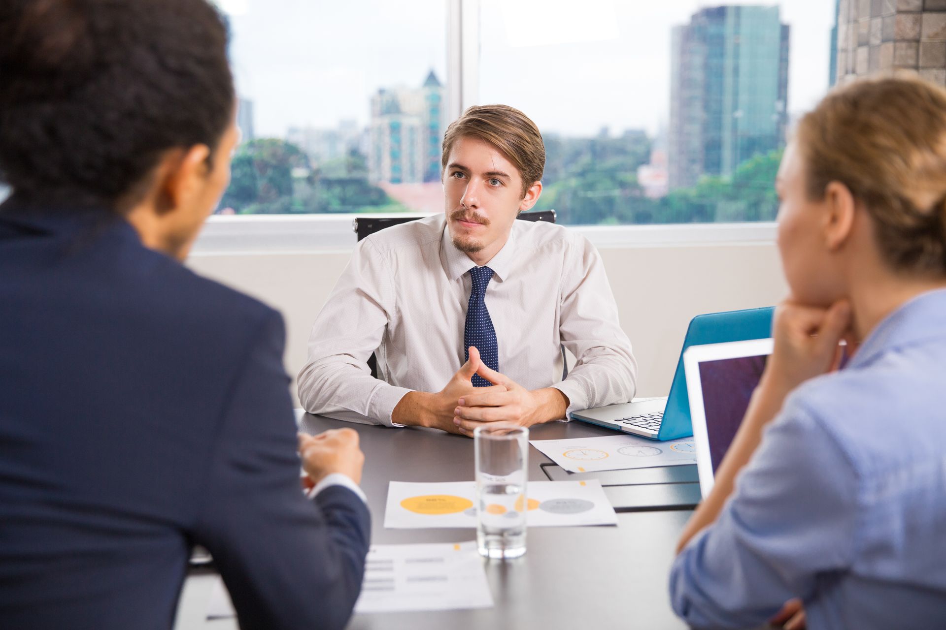 A business meeting in a bright office with a person speaking to two colleagues seated across a desk with charts.