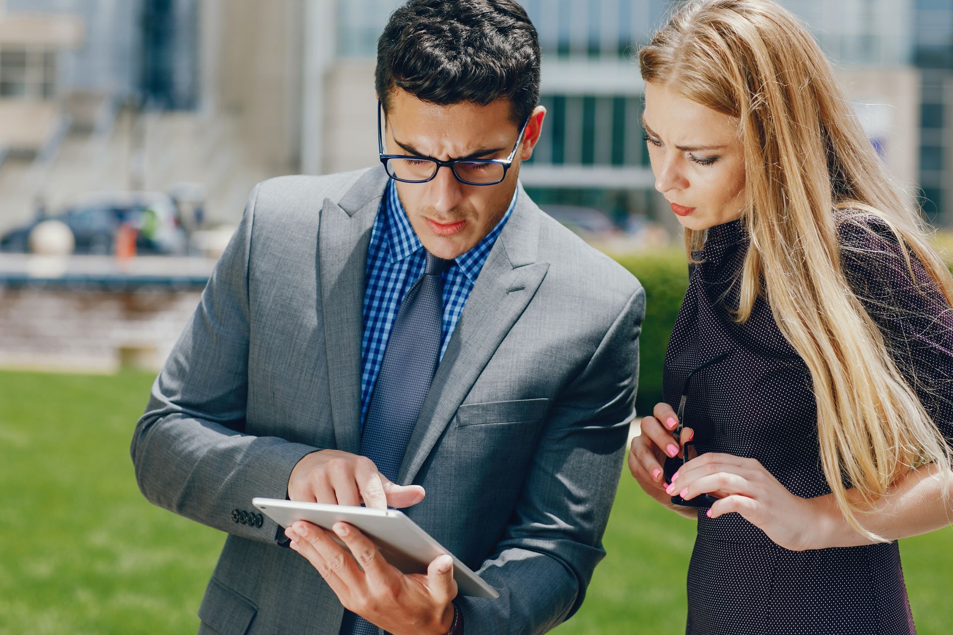 A professional man in a grey suit and a woman in a dark dress consult a tablet while standing outdoors near a building.