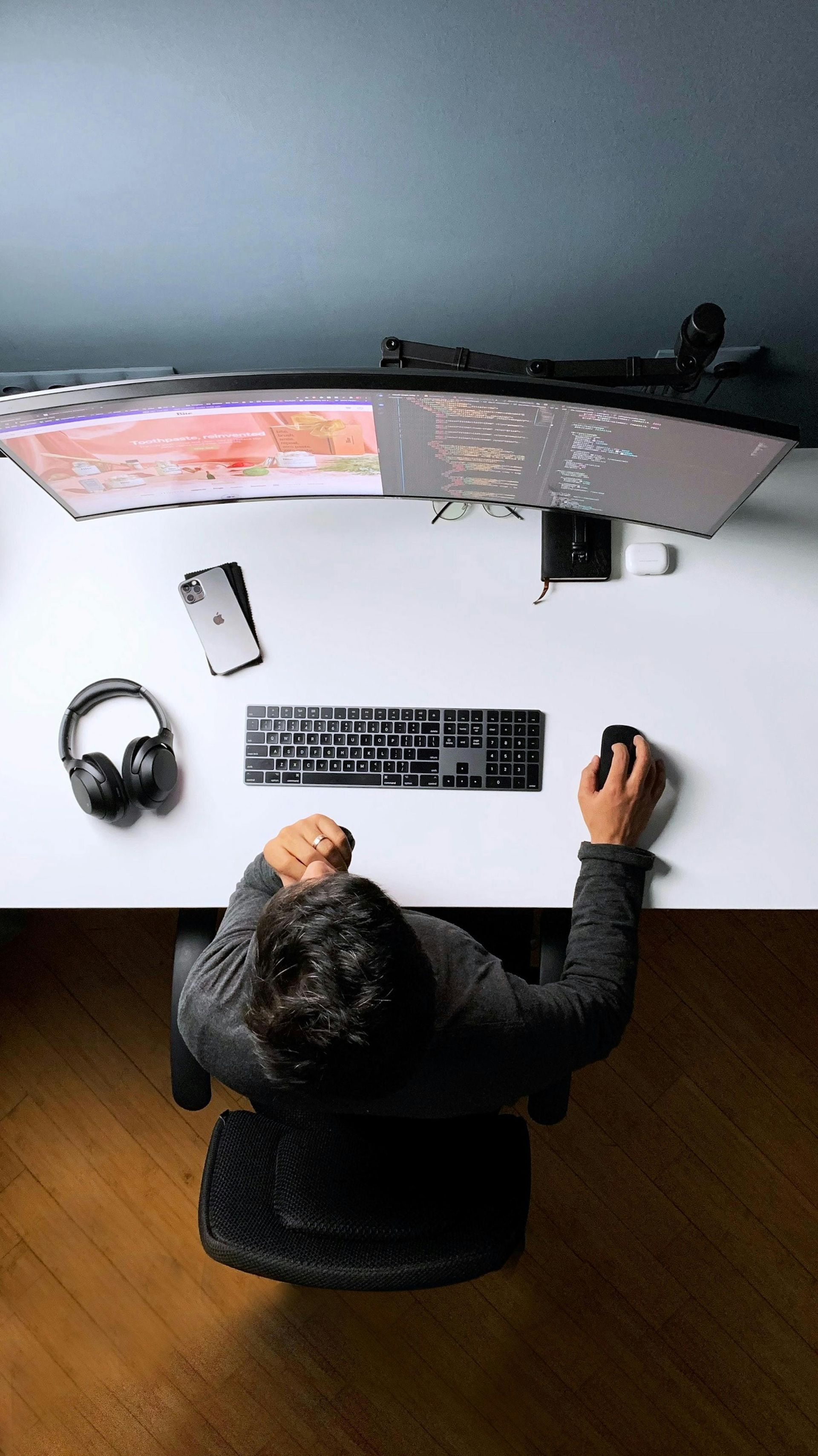 Top-down view of a person working at a white desk with a curved monitor, keyboard, mouse, headphones, and smartphone.