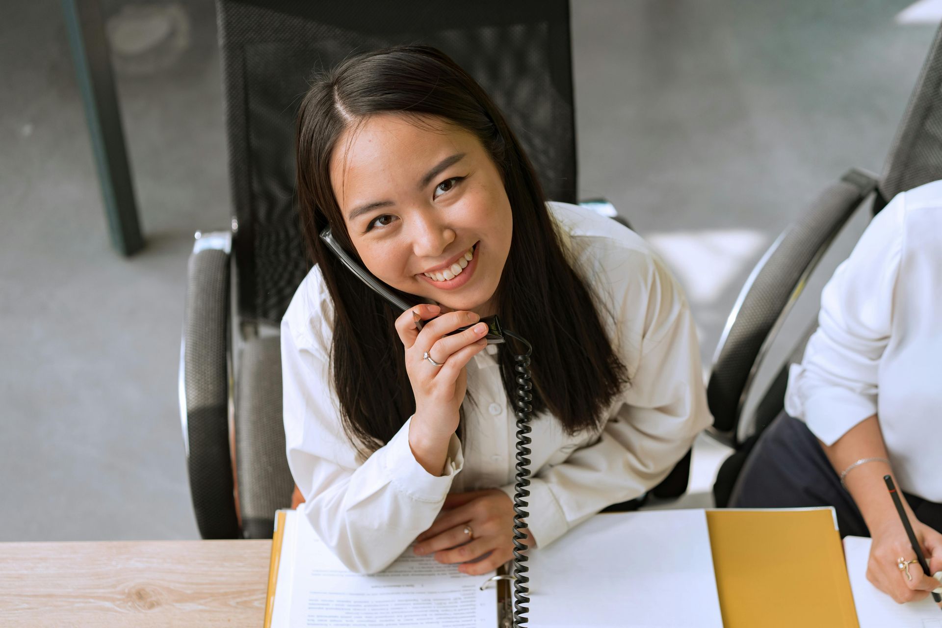 A smiling person sitting at a desk and holding a telephone handset to their ear while looking up at the camera.