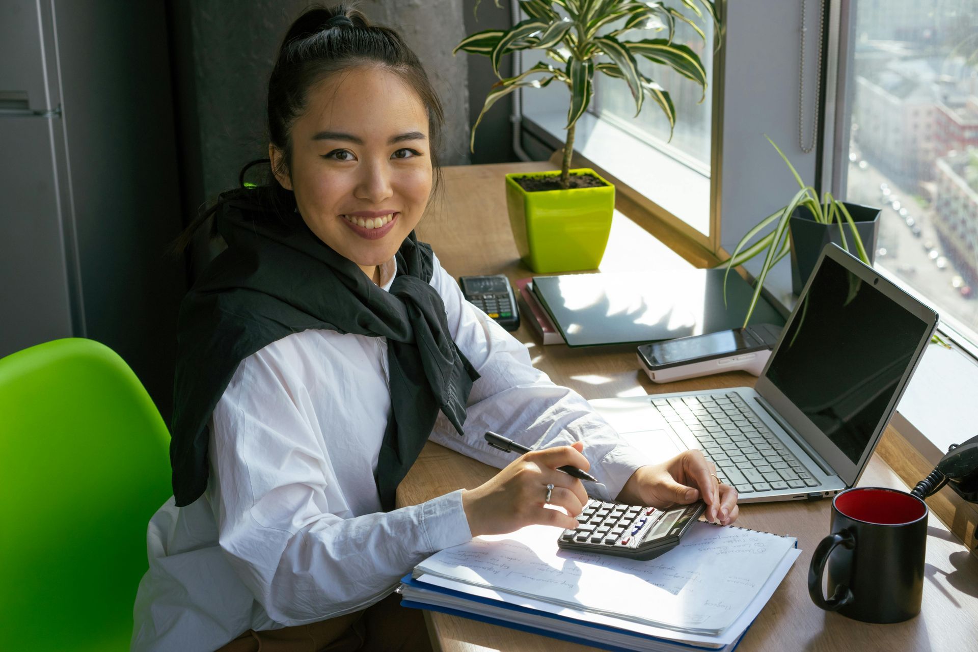 Woman smiles while working on a laptop, using a calculator at a desk by a window.