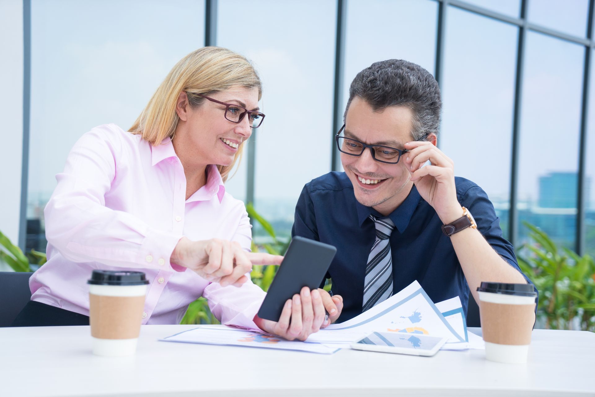 Two professionals in an office setting smile while reviewing information on a tablet, with coffee cups on the table.