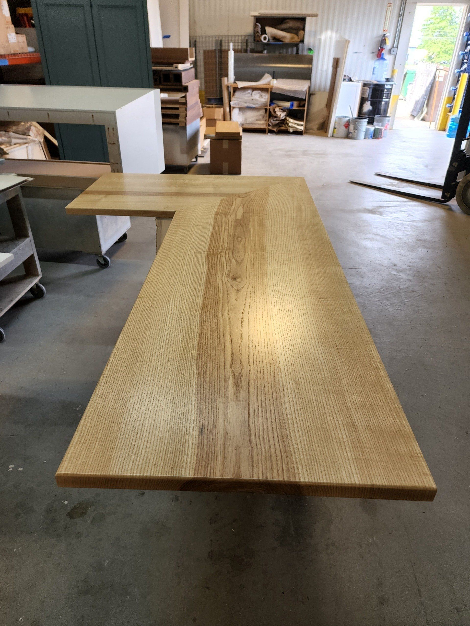 Large wooden L-shaped desk in a workshop setting. The desk has a light wood grain.