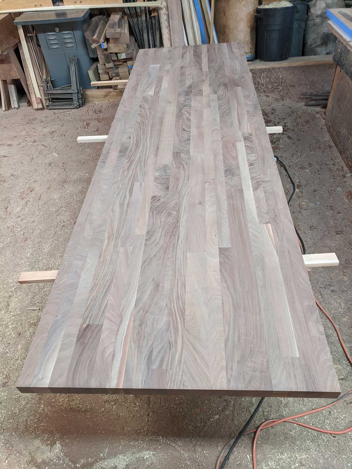 Long, rectangular wooden table top, light brown with grain, resting on supports in a workshop.