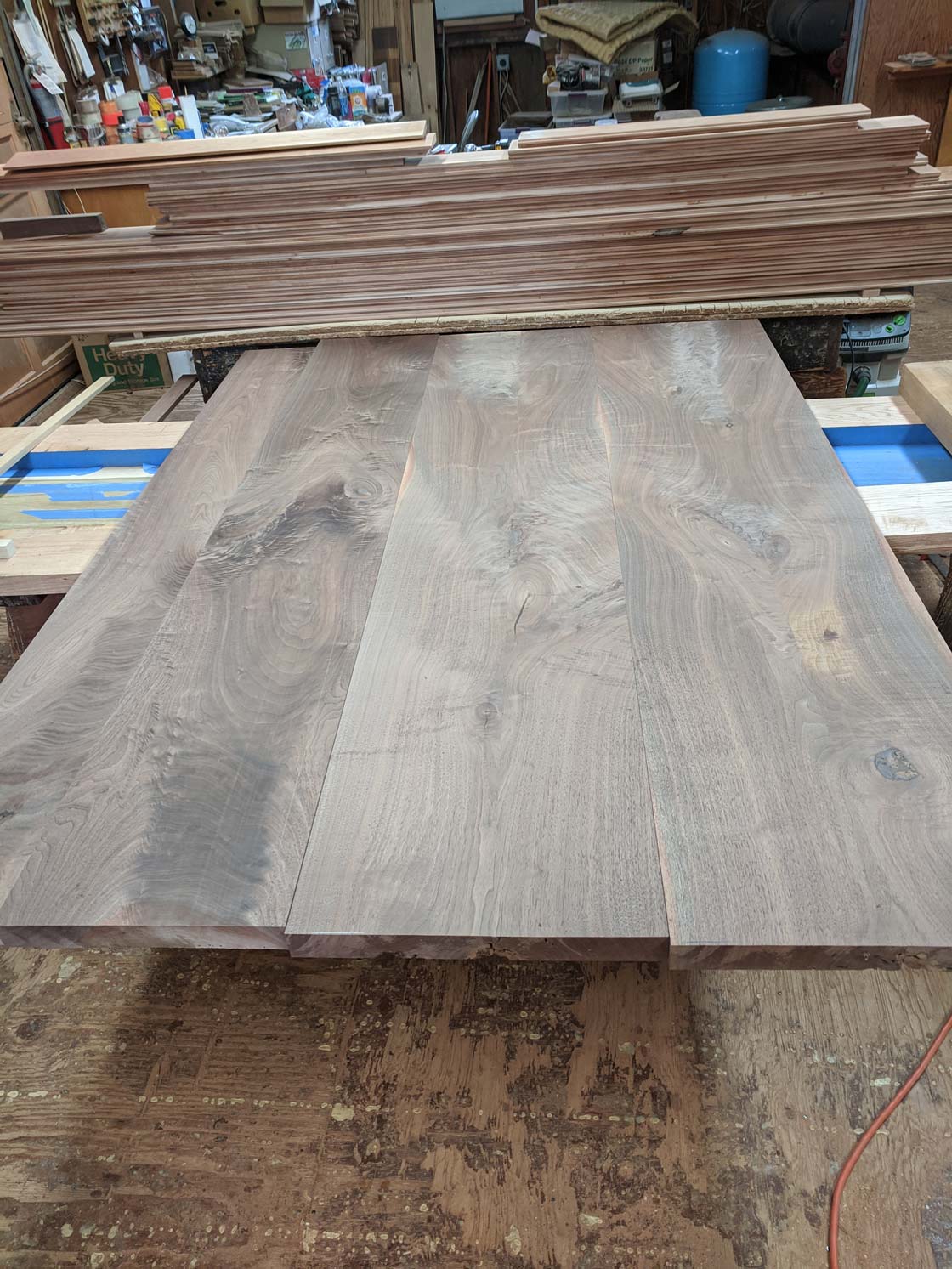 Wooden planks laid on a work surface, with more wood stacked in the background, workshop setting.