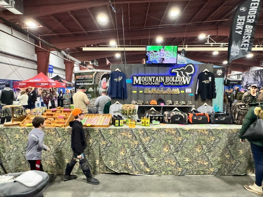 A vendor booth at an indoor event, with merchandise like shirts, hats, and bags. Camo tablecloth and sign reading 