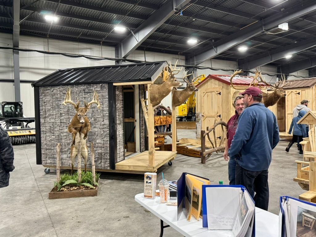 Hunting structures on display at a trade show. People look at various wooden shelters, some with mounted deer heads.