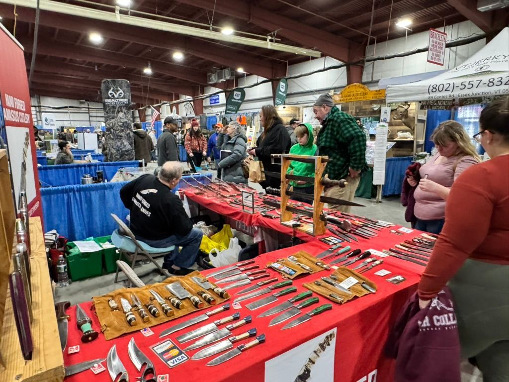 People browsing knives displayed on tables at an indoor event, with vendors and other booths visible.