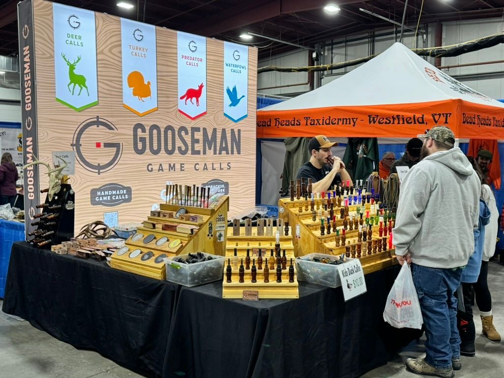 Gooseman Game Calls booth at a fair with hunting calls on display. Man talks to customers under a white tent.