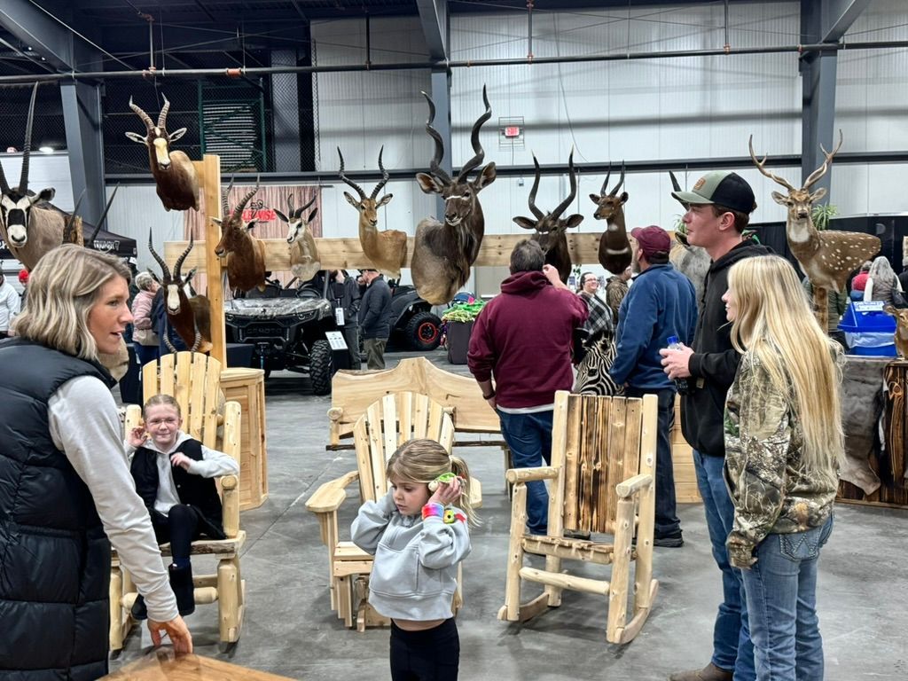 People view hunting trophies at an exhibition. Wooden chairs are in the foreground, taxidermied animals in background.