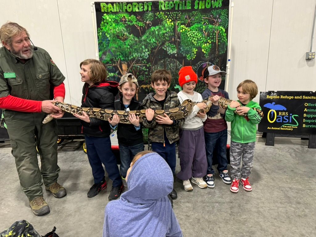 Man and children hold a large snake at a reptile show with a rainforest backdrop.