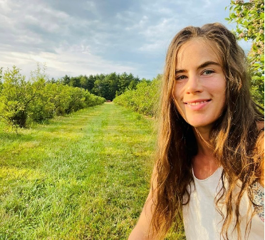 Woman smiles in a green field with rows of bushes, trees in the background, cloudy sky.
