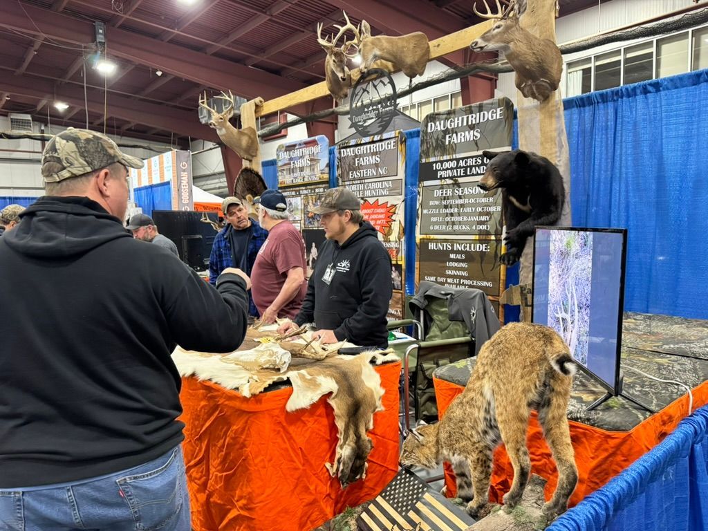 People browsing a taxidermy display at a trade show. Mounted animals, furs, and signs are visible.