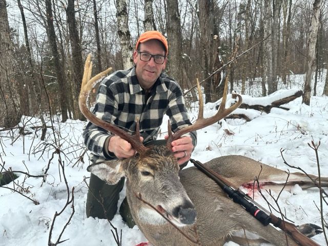 Man kneels in snow, holding deer antlers; rifle lies nearby. Winter forest background.