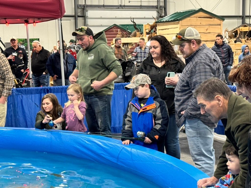 People fish at a small pool at an event. Others watch. A few kids are fishing, smiling. An indoor setting with booths.