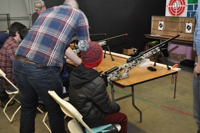 Person assisting a child with a rifle at a shooting range. Target and other rifles are in the background.