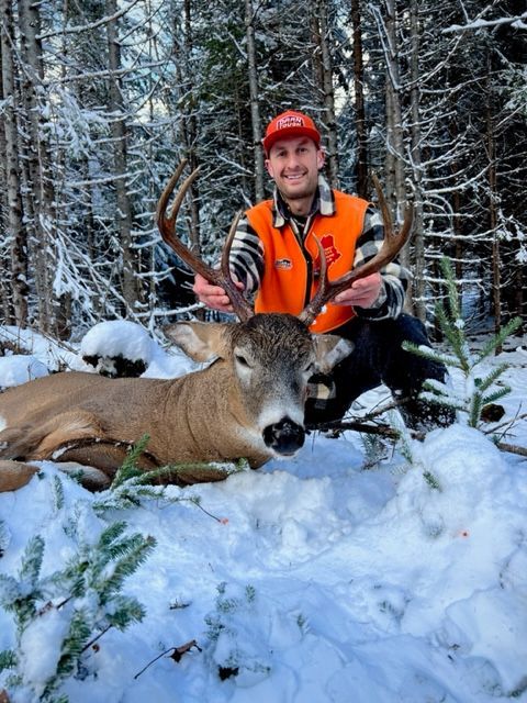 Hunter kneeling in snow, smiling, holding deer antlers next to a harvested deer. Forest background, orange vest.