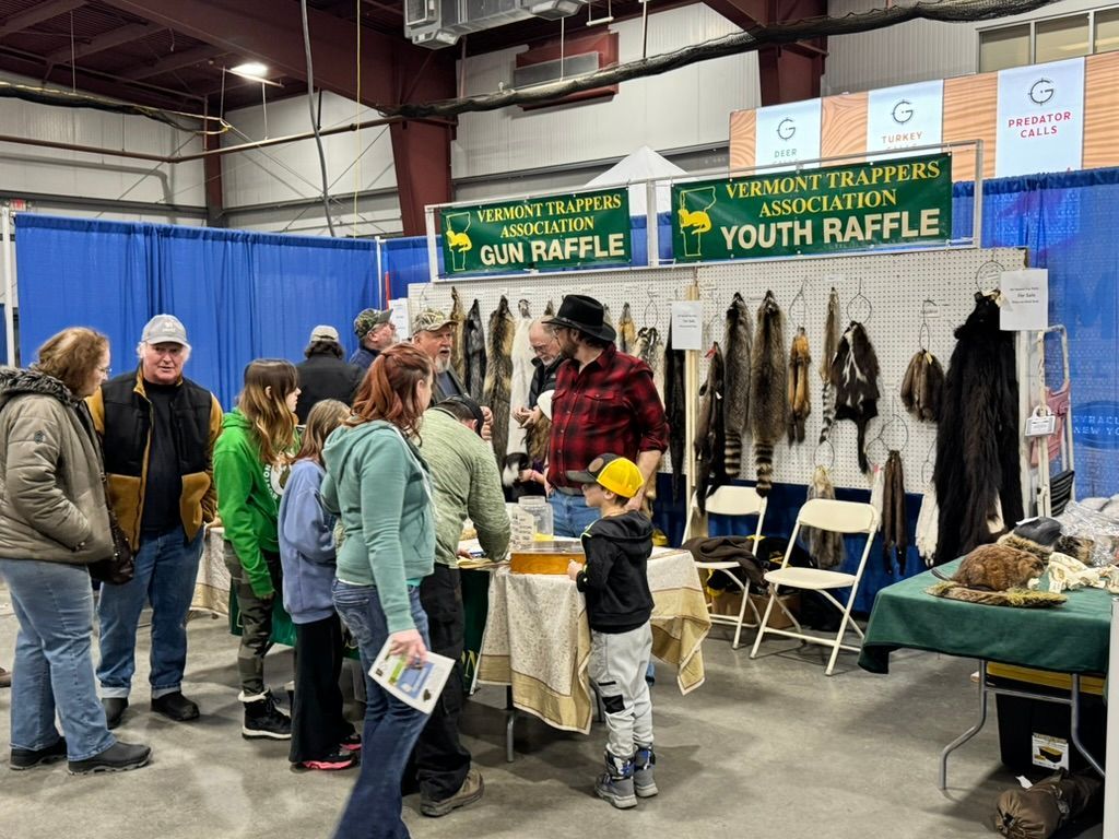 People at a trapping association booth. Pelts displayed, raffle signs, and attendees looking. Indoor setting.