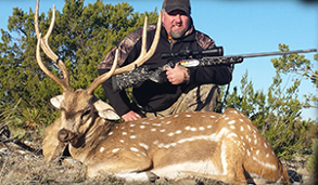 Man kneels beside a spotted deer he hunted, holding a rifle in a grassy, outdoor setting.
