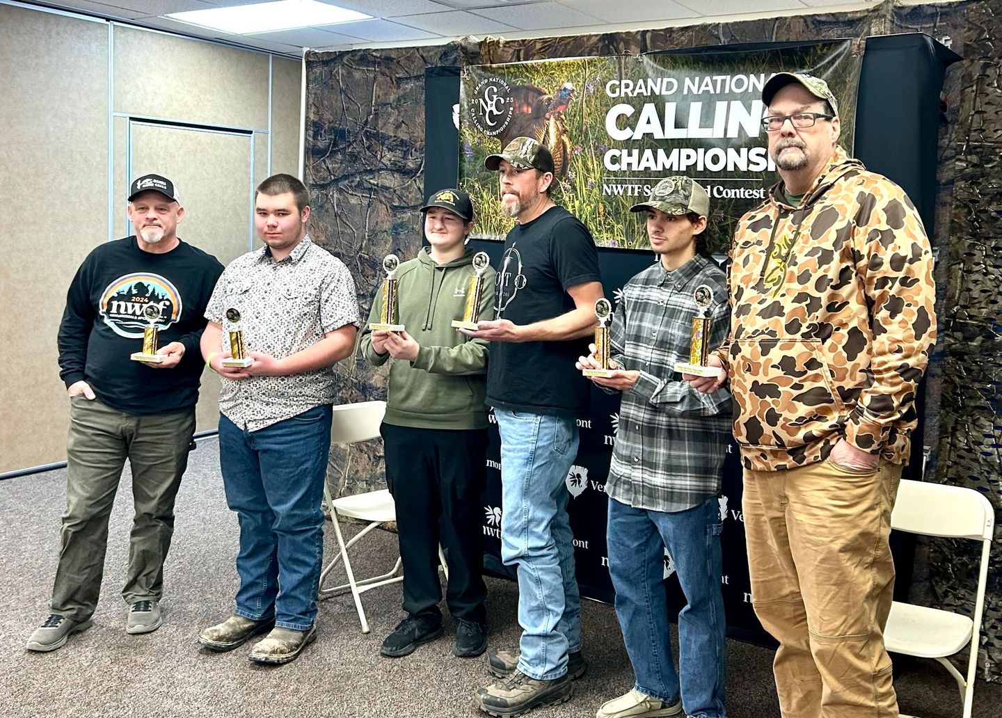 Men in casual attire holding trophies at a Grand National Calling Championship event. They are standing indoors.